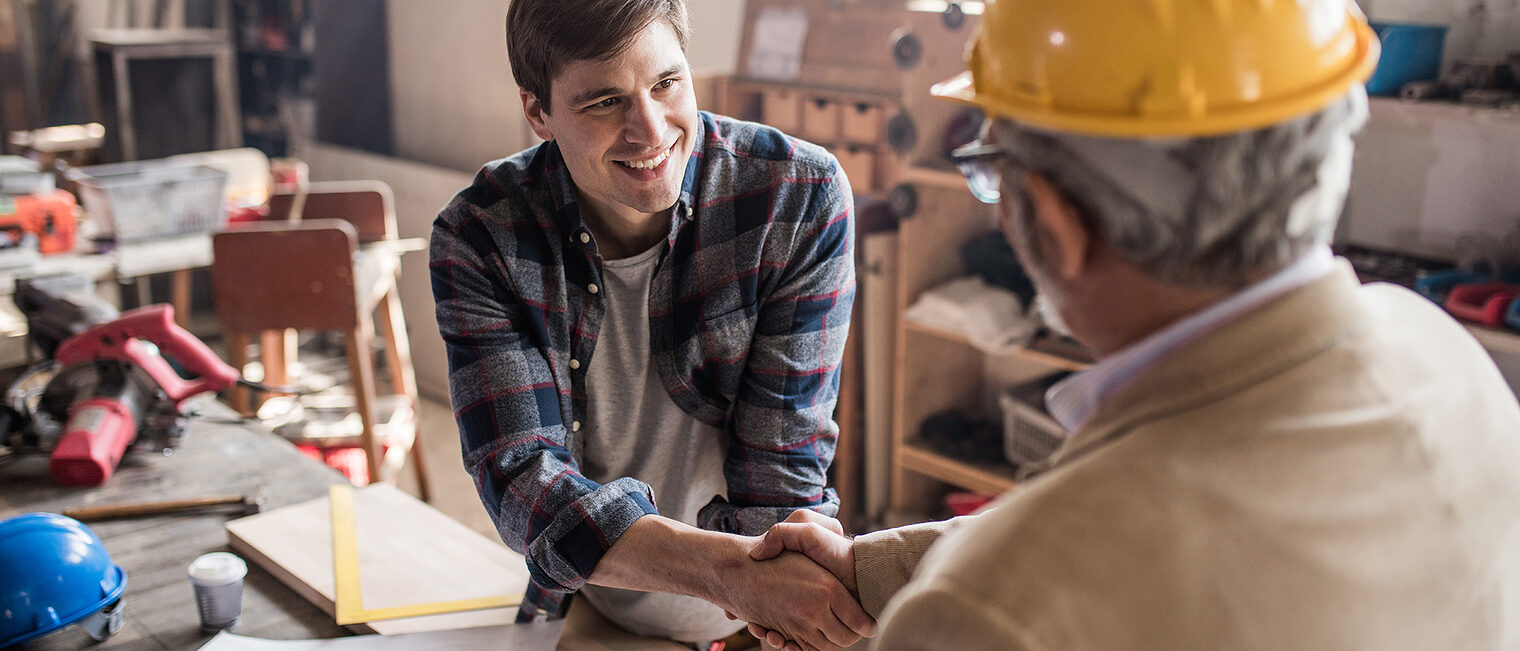 Young smiling carpenter shaking hand with quality control inspector in a workshop.