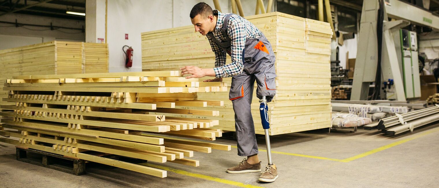 Ein junger Mann mit Beinprothese misst die Maße einer Holzlatte ab.