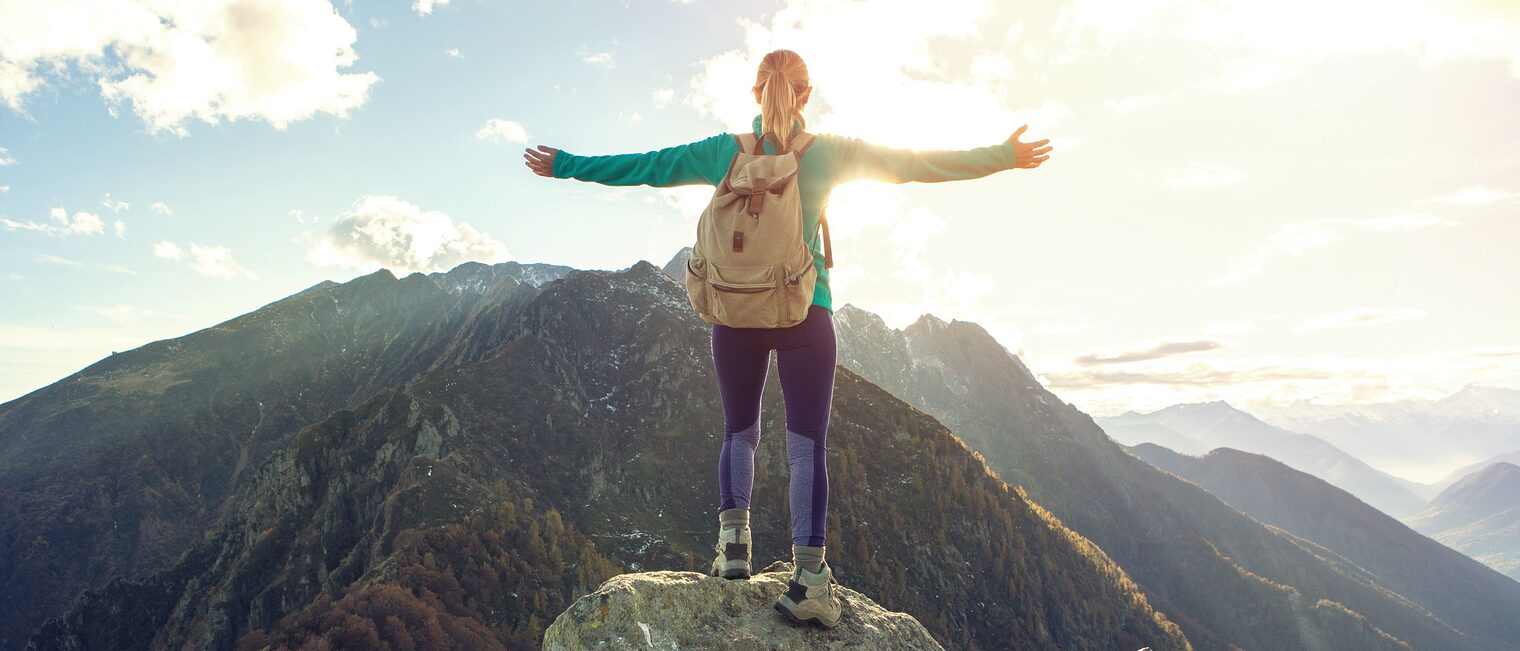 Eine junge Frau steht auf einem Berggipfel und streckt die Arme aus.