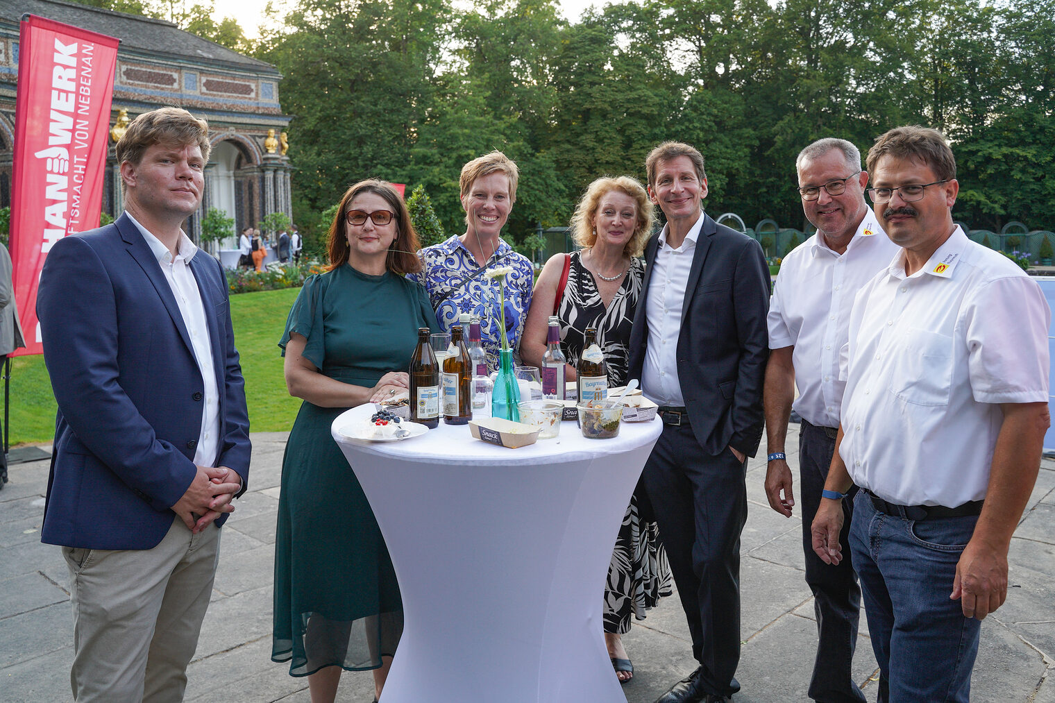 Gruppe von sieben Personen bei einem Stehempfang im Freien, alle lachen und genießen Getränke am Tisch.