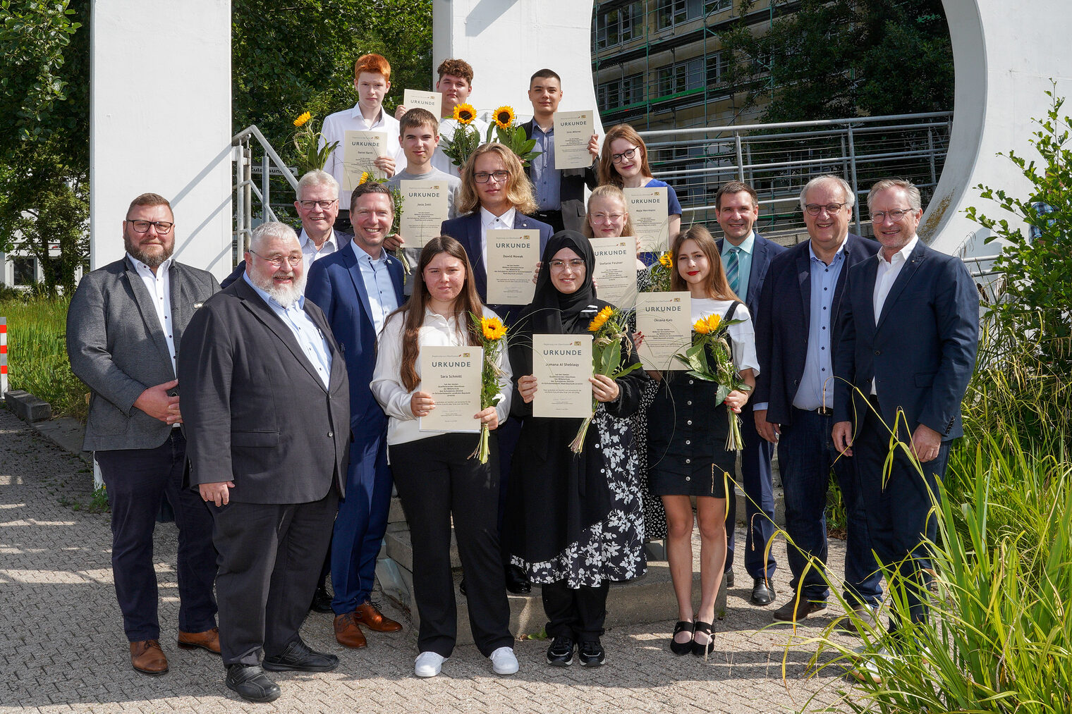 Gruppenfoto von Schülern mit Urkunden und Blumen bei der Bestenehrung 2024, umgeben von Lehrern und Vertretern, im Freien