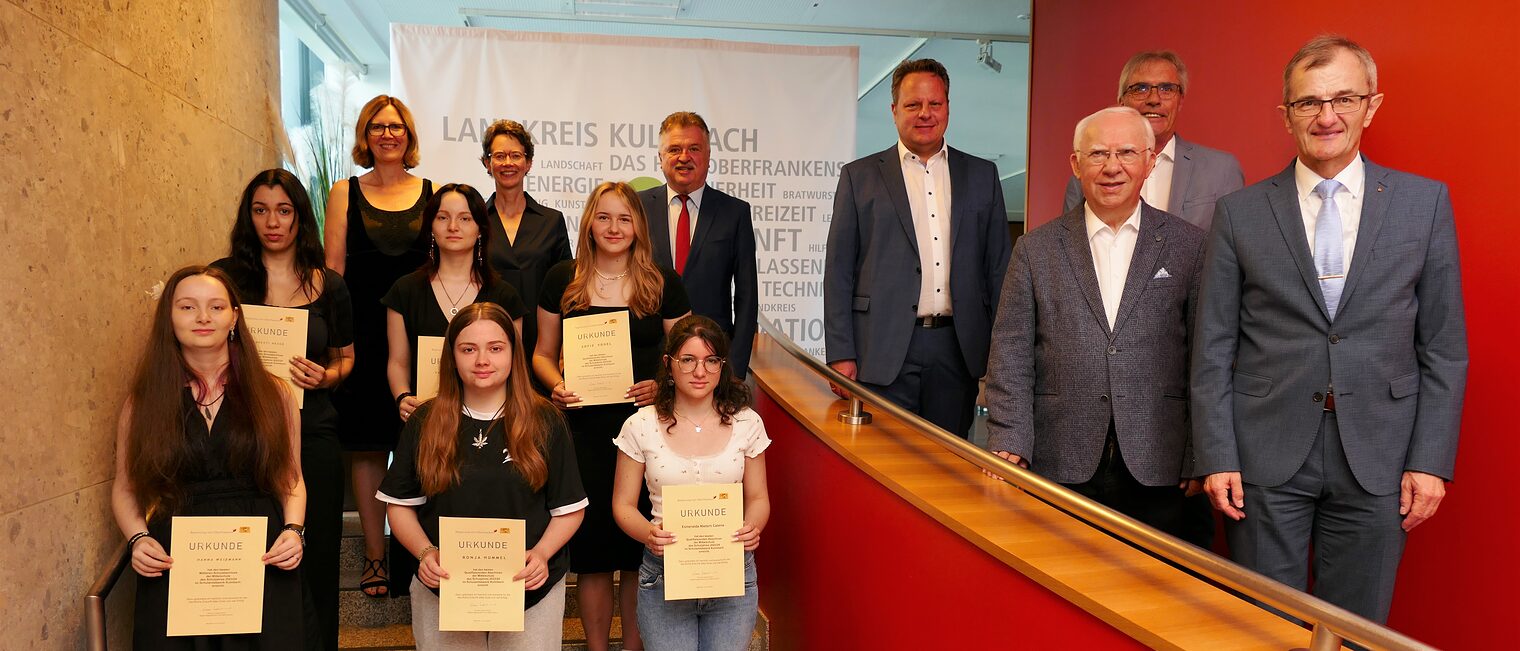 Gruppenfoto der besten Absolventinnen aus dem Schulamtsbezirk Kulmbach, die auf einer Treppe Urkunden halten, umgeben von Lehrern und Vertretern.