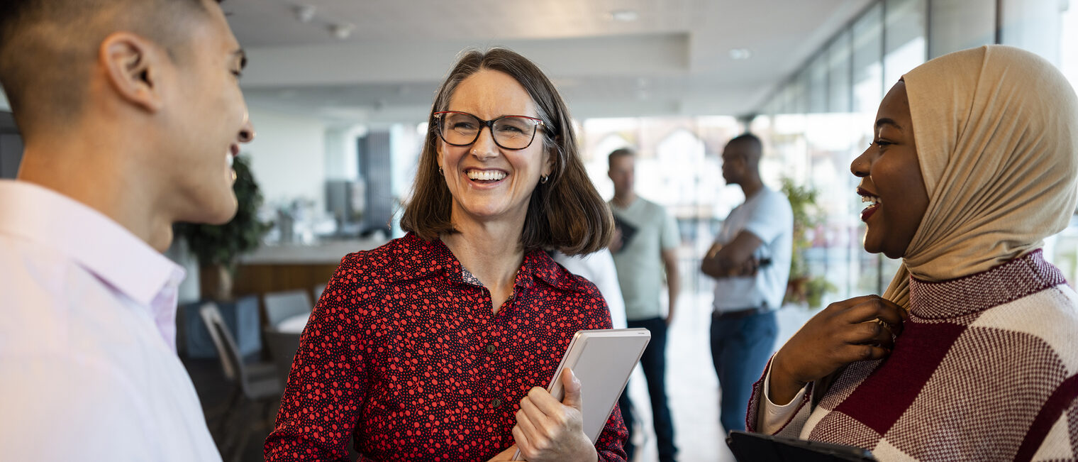 Drei Personen unterhalten sich lächelnd in einem modernen Büro, eine Frau hält ein Tablet, weitere Menschen im Hintergrund.