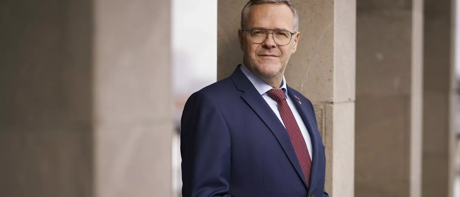 Jörg Dittrich, Präsident des ZDH im Anzug mit Brille und roter Krawatte steht vor einer Steinwand und blickt in die Kamera.