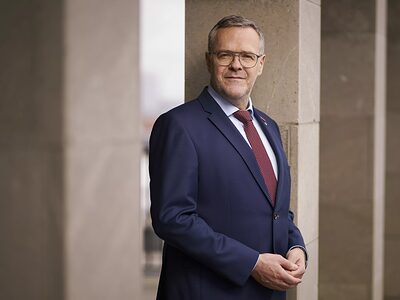 Präsident Dittrich im Anzug, roter Krawatte und Brille vor Steinwand. Jörg Dittrich, Präsident des ZDH im Anzug mit Brille und roter Krawatte steht vor einer Steinwand und blickt in die Kamera.