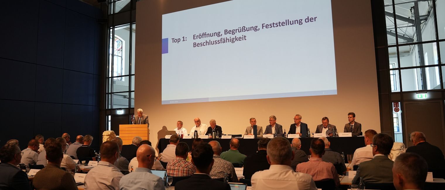 Der Vorstand und die Geschäftsführung der Handwerkskammer für Oberfranken sitzt an einem Tisch auf einem Podium vor der Vollversammlung.