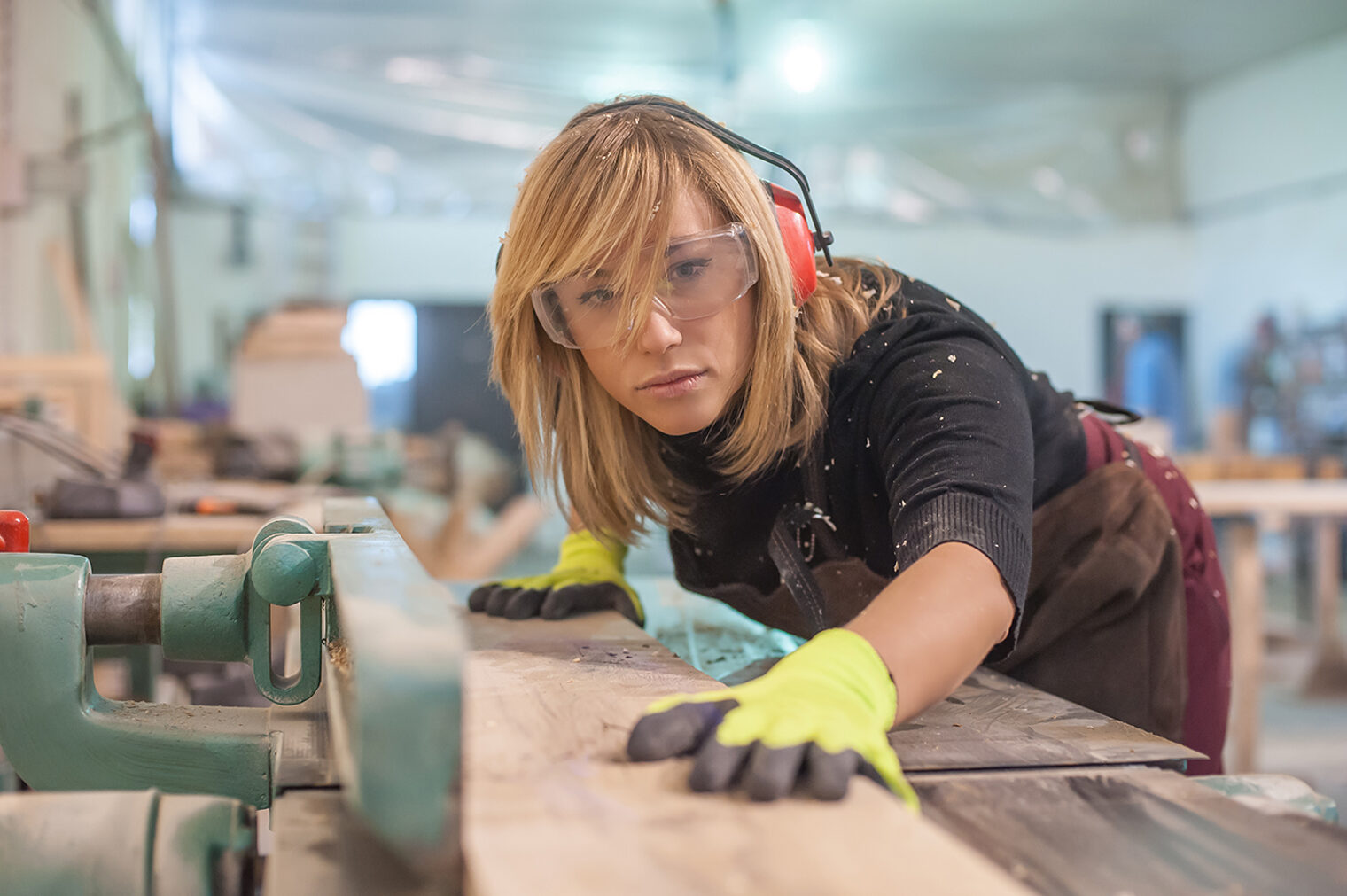 Eine Schreinerin mit Schutzbrille, Gehörschutz und Handschuhen schneidet ein Brett zu. 