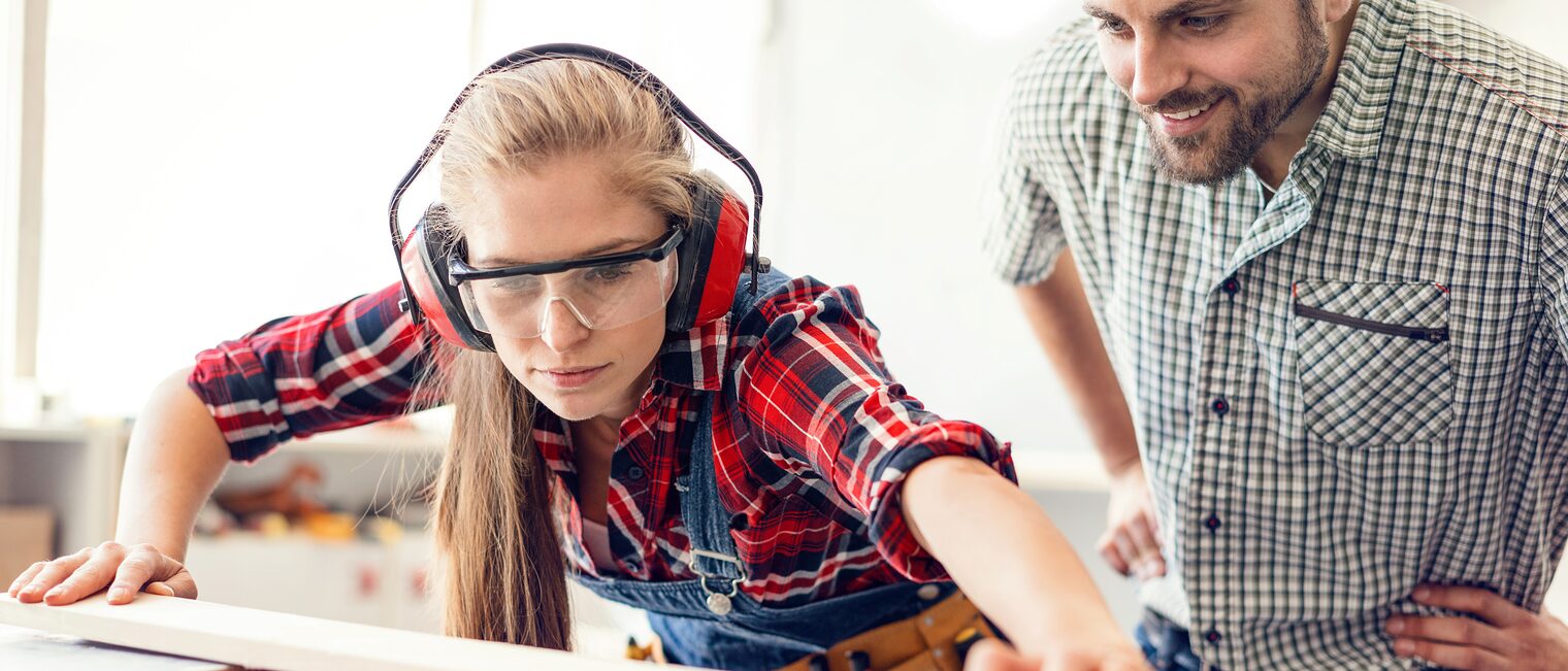 Eine junge Frau mit Gehörschutz und Schutzbrille sägt unter Aufsicht ein Holzbrett zurecht. 