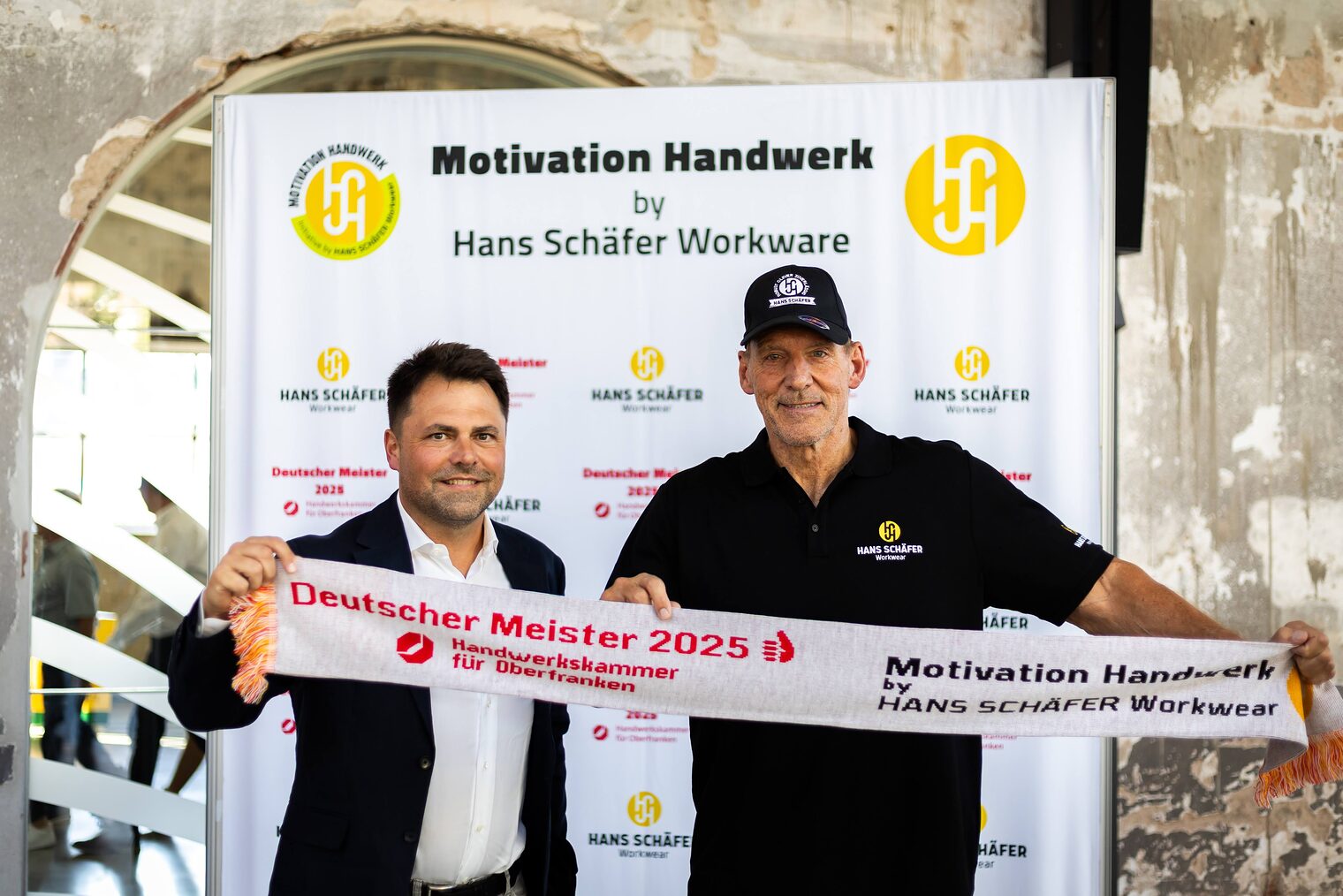 Andreas Grewe und Ralf Moeller posieren vor einer Fotowand mit einem Meisterschal.