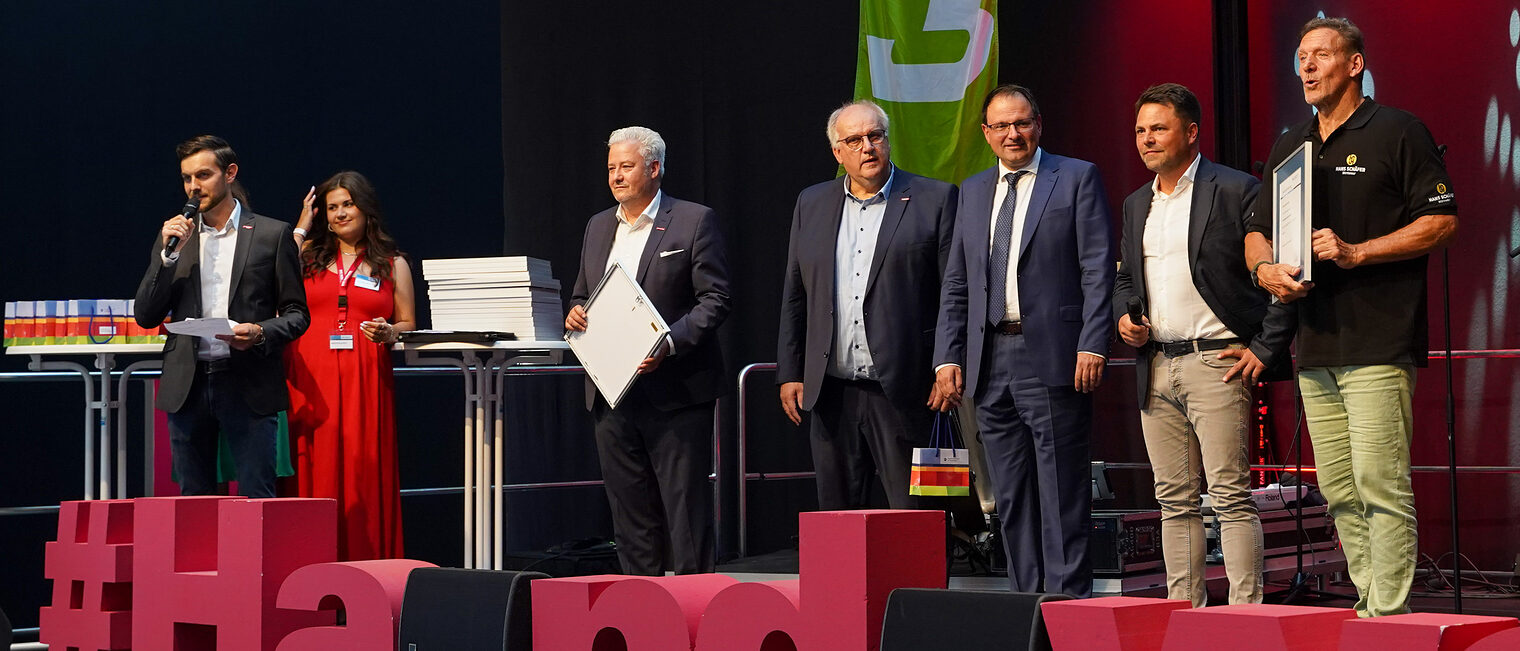 Sven Neukamm spricht auf der Bühne in ein Mikrofon während Matthias Graßmann, Reinhard Bauer, Martin Schöffel, Andreas Grewe und Ralf Moeller neben ihm stehen.