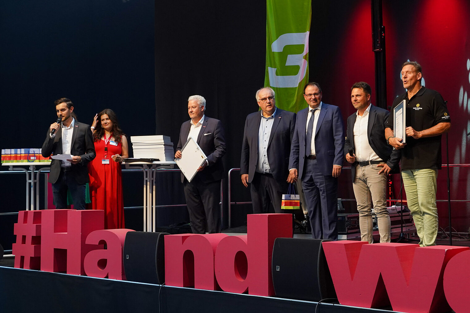 Sven Neukamm spricht auf der Bühne in ein Mikrofon während Matthias Graßmann, Reinhard Bauer, Martin Schöffel, Andreas Grewe und Ralf Moeller neben ihm stehen. 