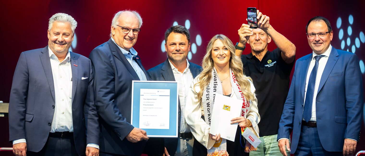 Friseurmeisterin Jacqueline Koziol präsentiert ihre Bestenurkunde mit Matthias Graßmann, Reinhard Bauer, Andreas Grewe, Ralf Moeller und Martin Schöffel.