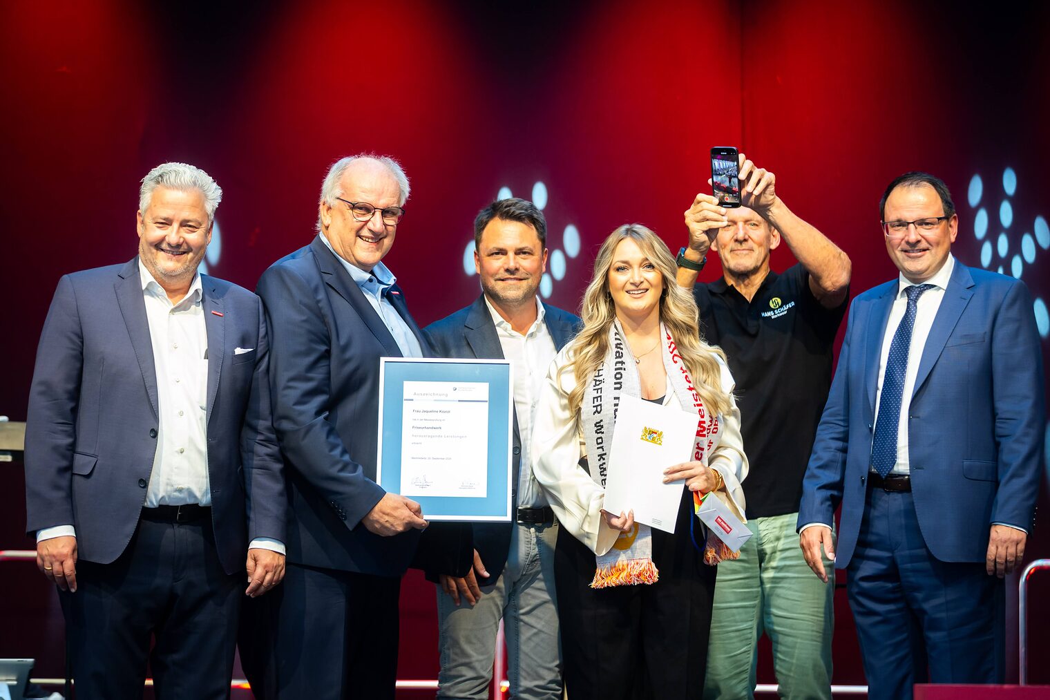Friseurmeisterin Jacqueline Koziol präsentiert ihre Bestenurkunde mit Matthias Graßmann, Reinhard Bauer, Andreas Grewe, Ralf Moeller und Martin Schöffel.