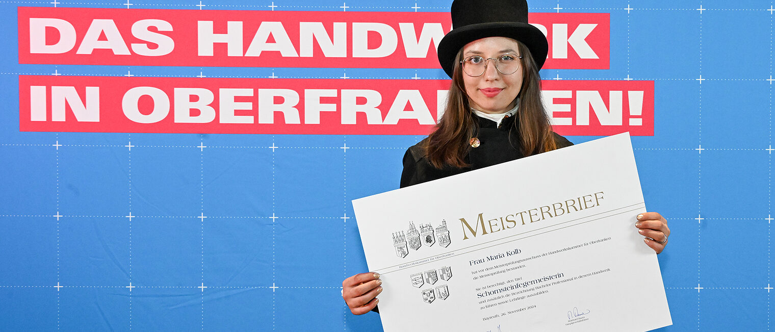 Schornsteinfegermeisterin Maria Kolb posiert mit ihrem Meisterbrief vor einer Fotowand.