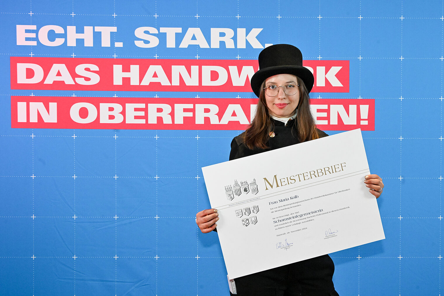 Schornsteinfegermeisterin Maria Kolb posiert mit ihrem Meisterbrief vor einer Fotowand. 