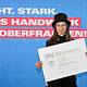 Schornsteinfegermeisterin Maria Kolb posiert mit ihrem Meisterbrief vor einer Fotowand. 