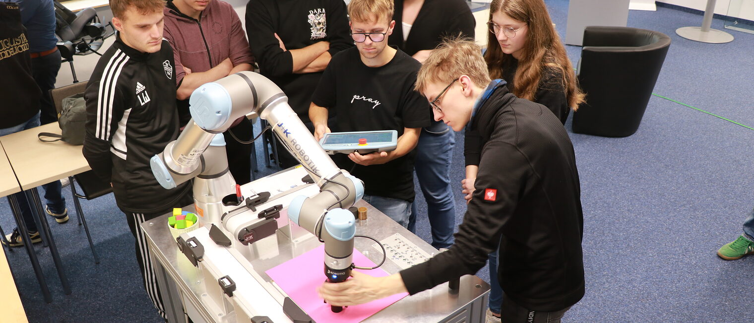 Eine Gruppe Teilnehmer und Teilnehmerinnen sieht zu wie ein Teilnehmer den Roboterarm über einem Testwürfel in Stellung bringt.