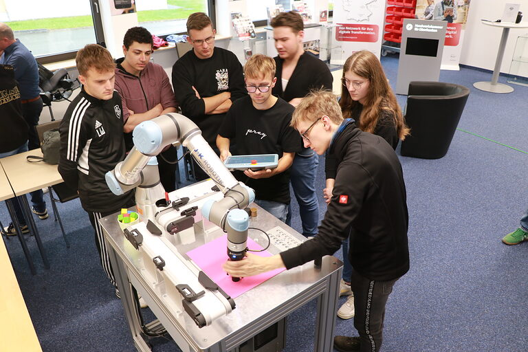 Eine Gruppe Teilnehmer und Teilnehmerinnen sieht zu wie ein Teilnehmer den Roboterarm über einem Testwürfel in Stellung bringt.