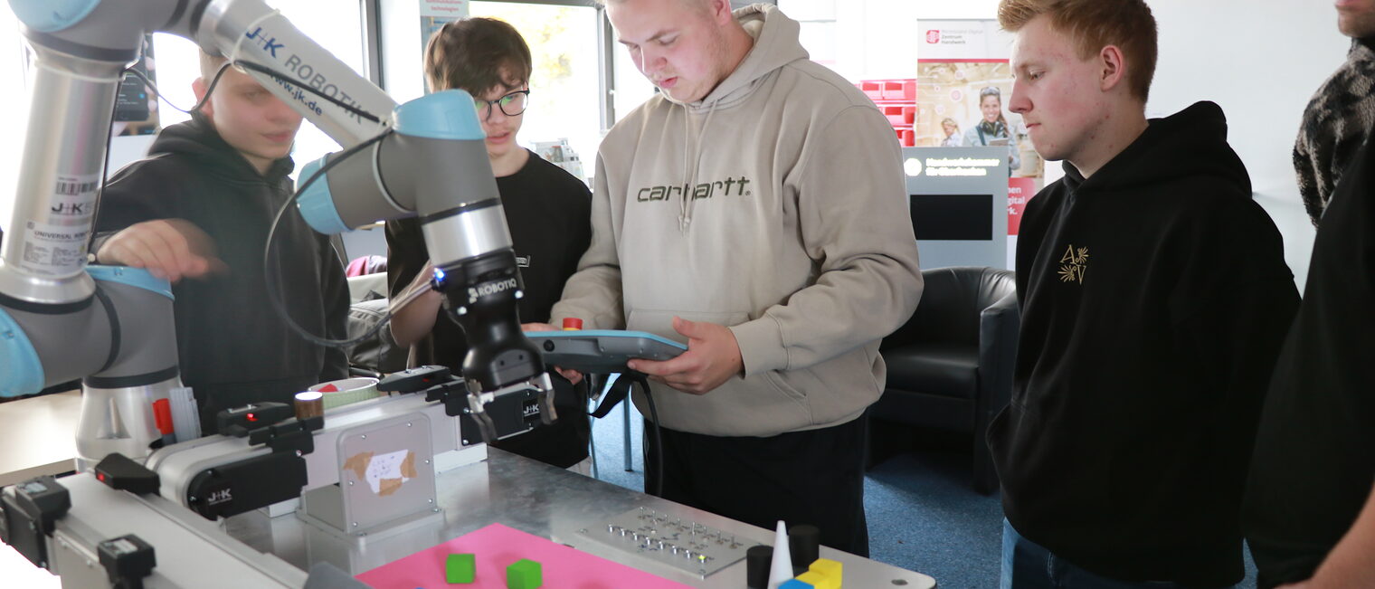 Eine Gruppe Auszubildender bedient am Tablet einen Roboterarm vor einem Untergrund mit Testwürfeln.