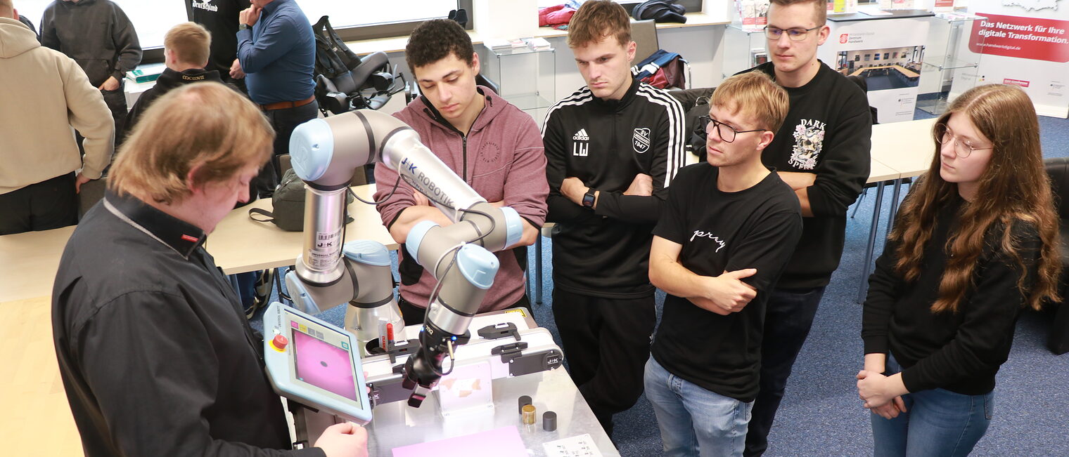 KI-Trainer Robert Falkenstein zeigt den Auszubildenden neben dem Roboterarm, wie sie Einstellungen am Tablet vornehmen.