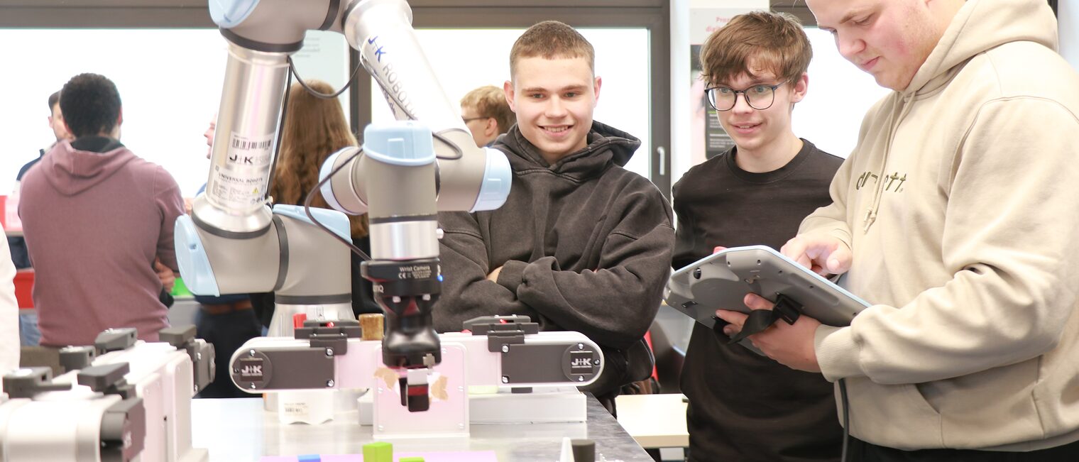 Ein Auszubildender nimmt Einstellungen am Tablet neben dem Roboterarm vor, während andere Auszubildende ihm mit begeisterten Gesichtsausdrücken zusehen.