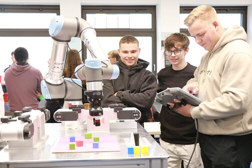 Ein Auszubildender nimmt Einstellungen am Tablet neben dem Roboterarm vor, während andere Auszubildende ihm mit begeisterten Gesichtsausdrücken zusehen.