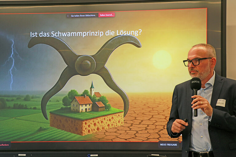 Professor Günter Müller-Czygan spricht neben einer Illustration der Schwammstadt mit einer Stadt im Klammergriff und Schwammgrundlage ins Mikrofon.