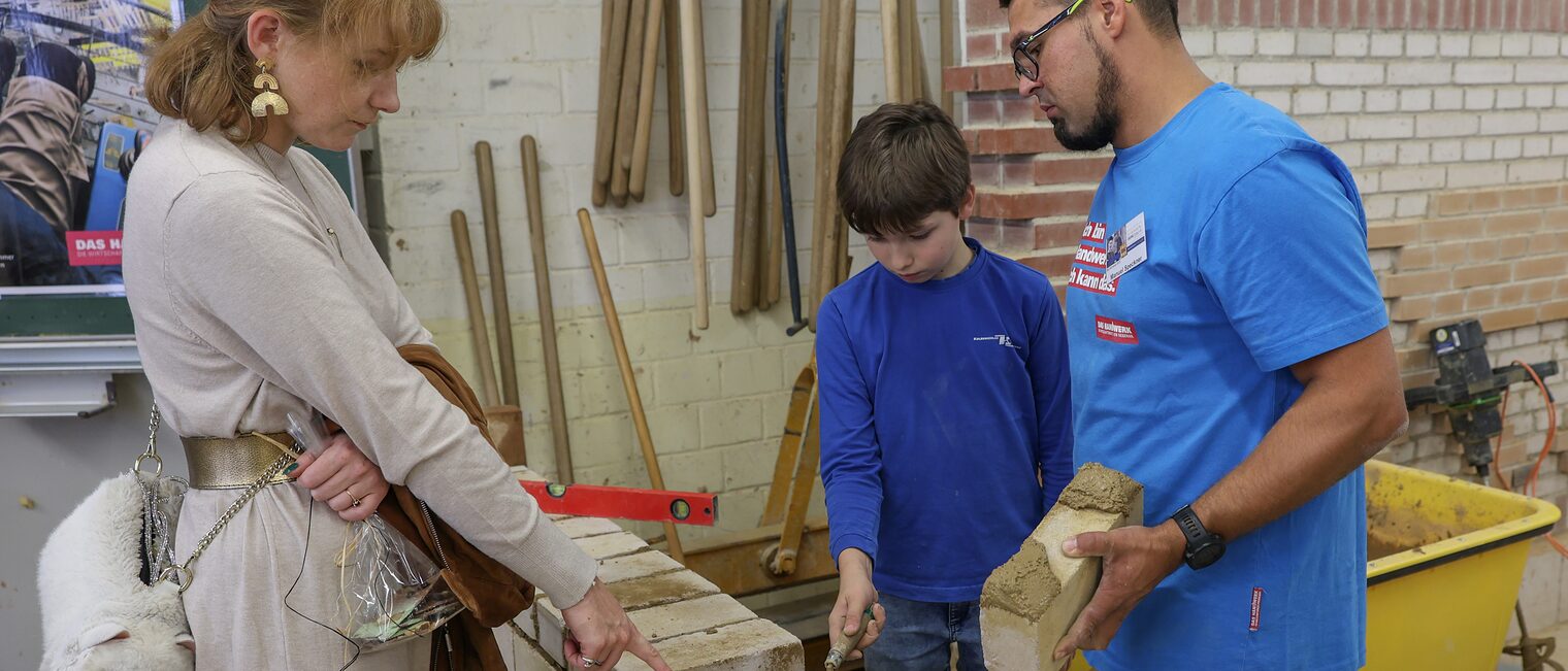 Eine Mutter beobachtet ihren Sohn, der unter Anleitung Steine auf eine Mauer setzt.
