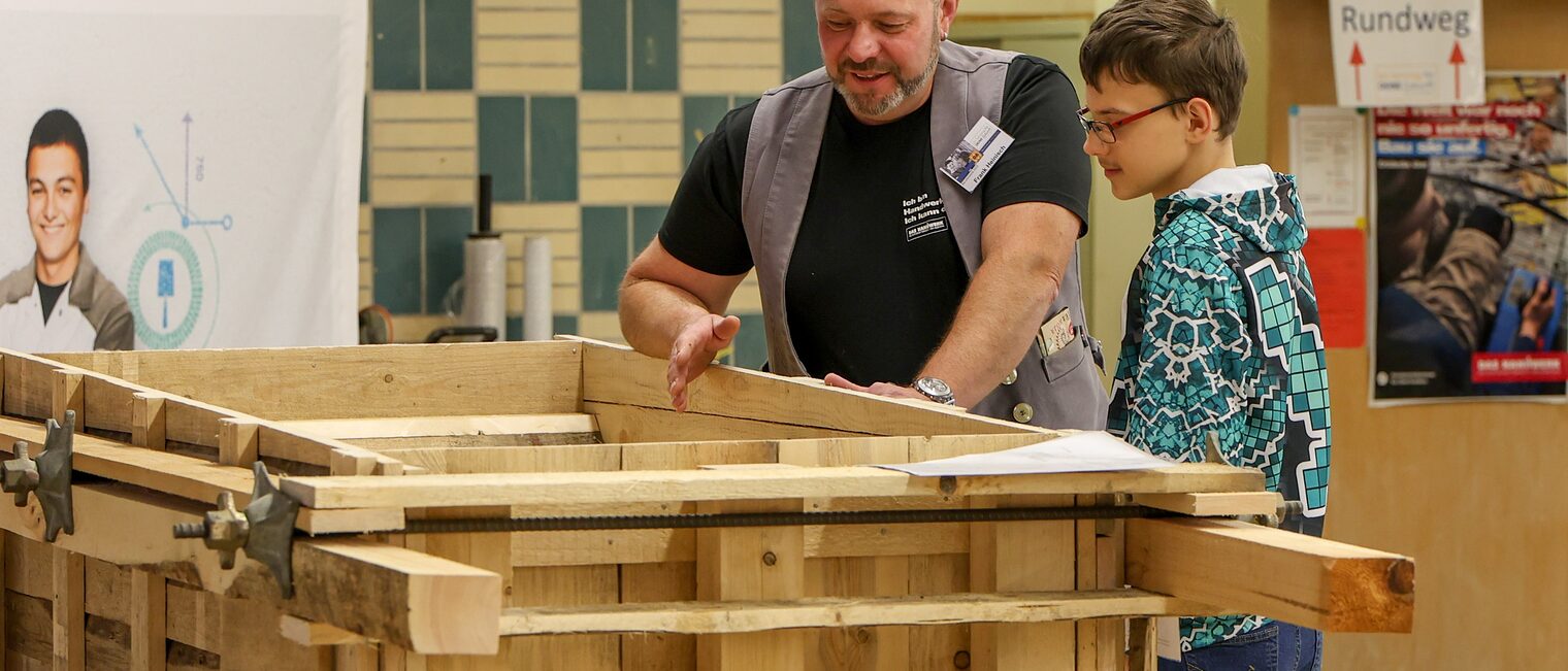 Ein Ausbildungsmeister zeigt einem Jugendlichen eine Holzkonstruktion.
