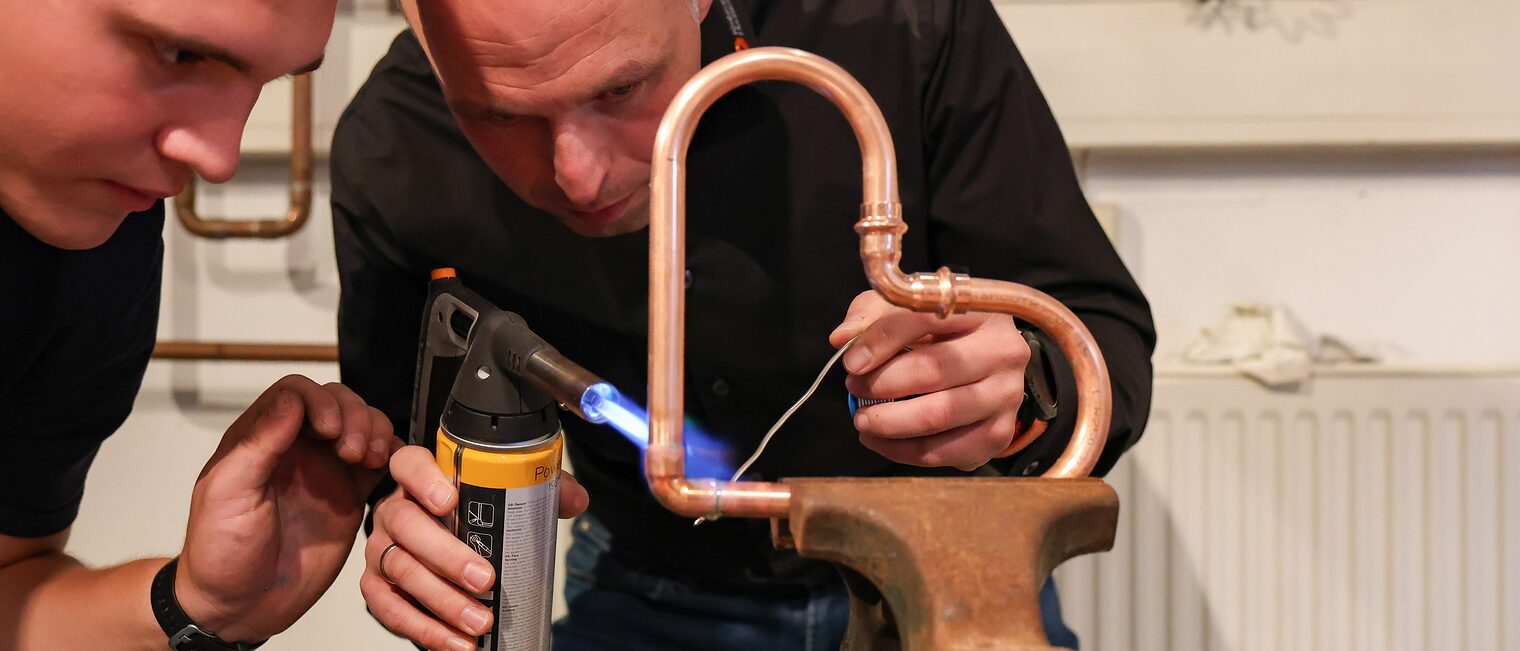 Ein Ehrengast sieht zu, wie ein Auszubildender ein Herz auf Kupferrohren schweißt.