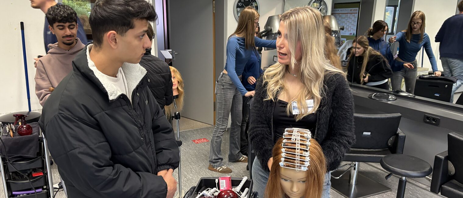 Friseurmeisterin Fenja Garrett zeigt einen Jugendlichen an einer Modellpuppe, wie er Locken wickelt.