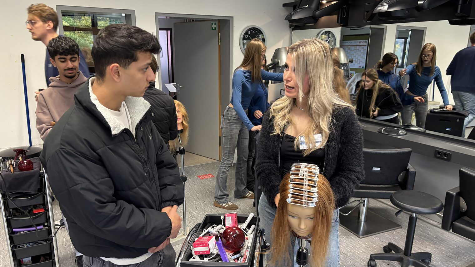 Friseurmeisterin Fenja Garrett zeigt einen Jugendlichen an einer Modellpuppe, wie er Locken wickelt.