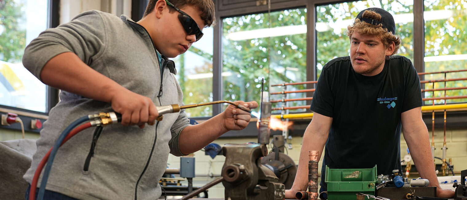 Ein Jugendlicher schweißt unter Aufsicht zwei Metallrohre zusammen.