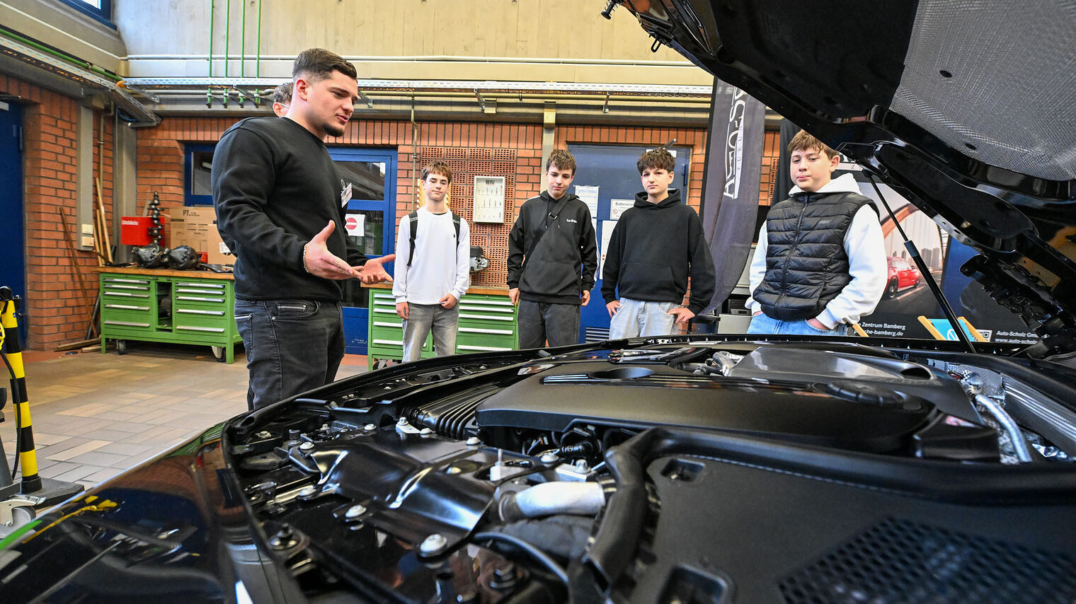 Ein Ausbildender zeigt Jugendlichen den Motorraum eines Fahrzeuges.