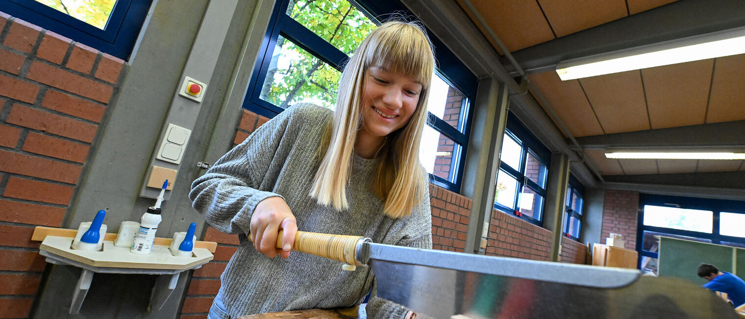 Eine Jugendliche sägt mit einem Schwertähnlichen Gegenstand ein Holzstück.