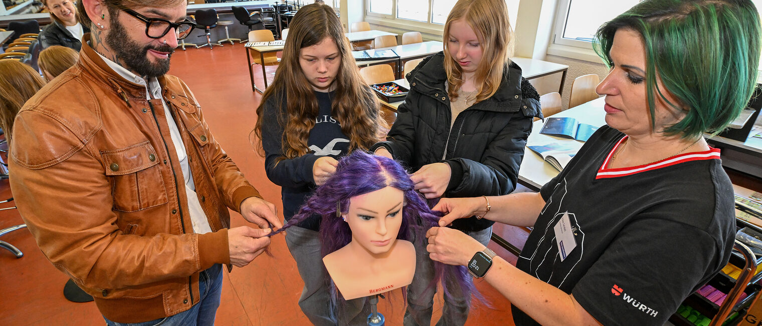 Eine Friseurmeisterin kämmt mit zwei Jugendlichen die Haare einer Modellpuppe.
