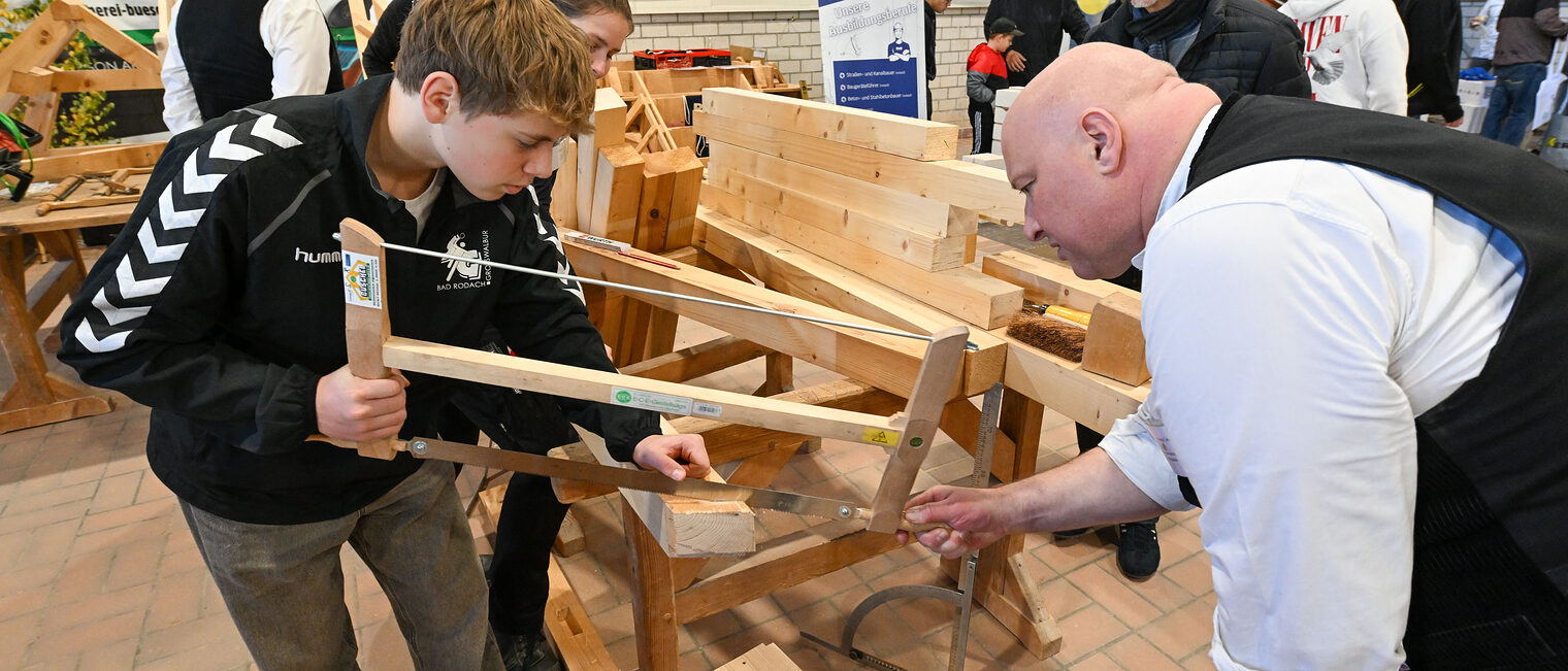 Ein Jugendlicher sägt unter Aufsicht ein Holzstück von einem Holzbalken.