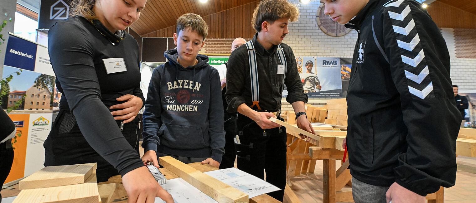 Eine Auszubildende zeigt Jugendlichen einen Bauplan auf einer Holzkonstruktion.