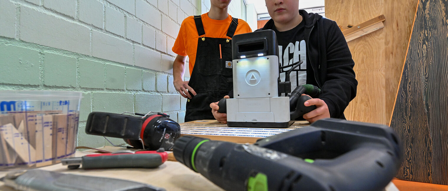 Ein Jugendlicher fräst unter Beaufsichtigung etwas in ein Holzstück.