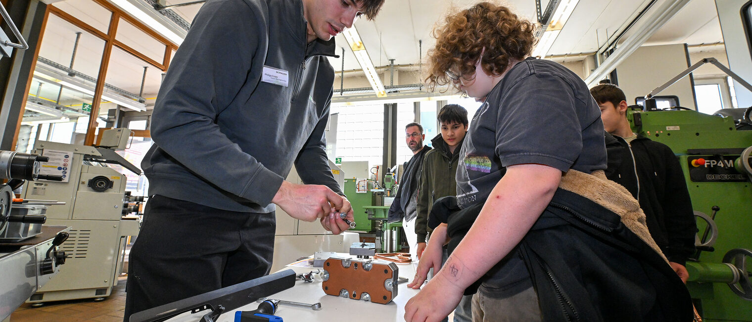 Ein Auszubildender zeigt einer Gruppe Jugendlicher Metallgewinde.