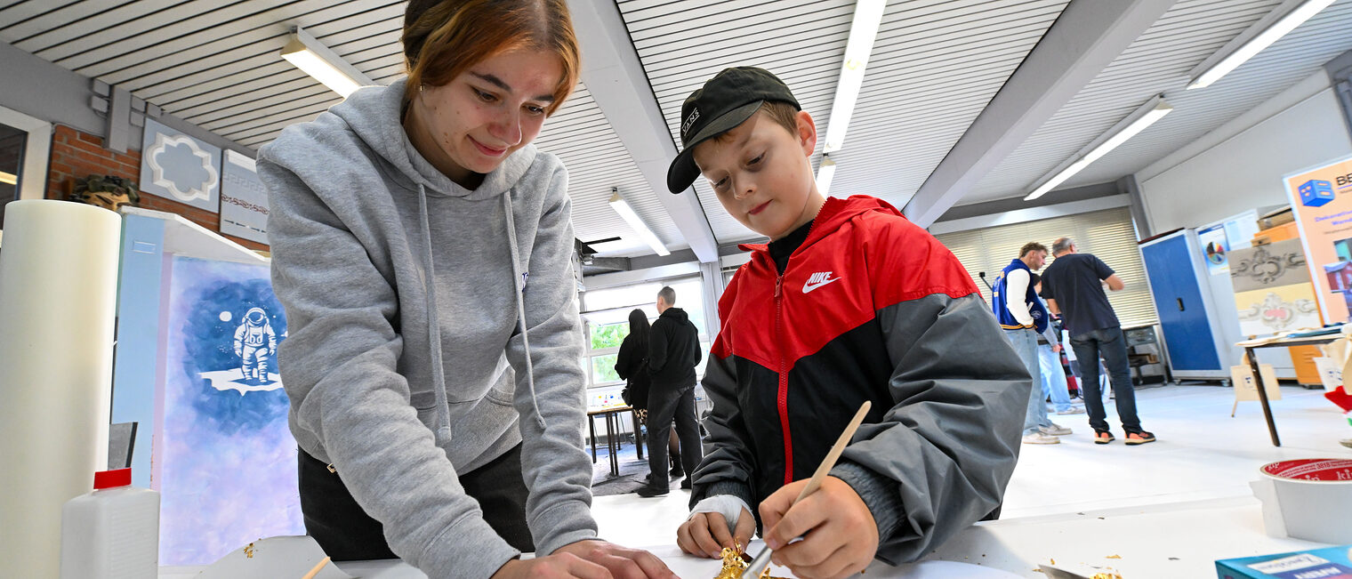 Ein Jugendlicher trägt mit einem Pinsel Goldfolie auf ein Blatt Papier auf, während eine Jugendliche es festhält. 