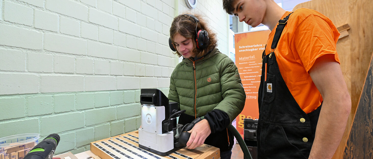 Ein Jugendlicher fräst unter Beaufsichtigung etwas in ein Holzstück.