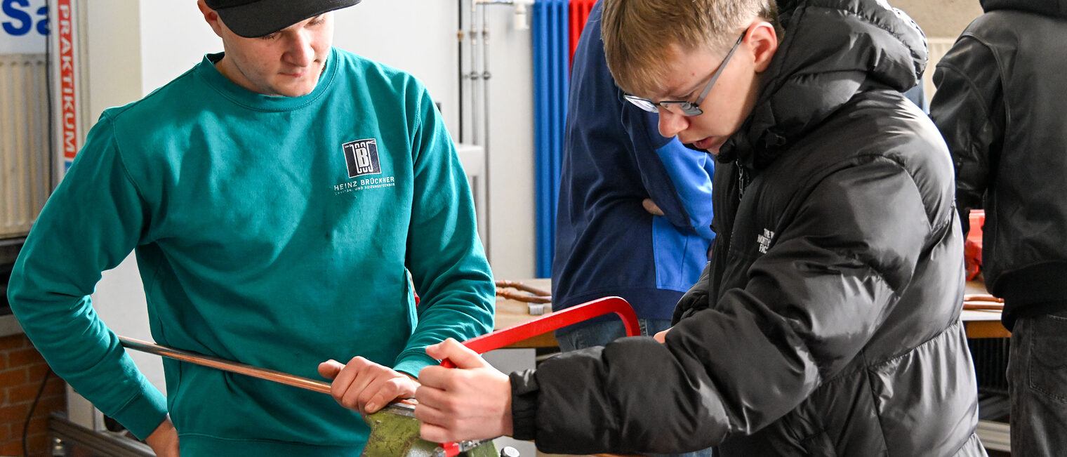 Ein Jugendlicher sägt unter Beaufsichtigung ein Metallrohr in zwei Teile. 