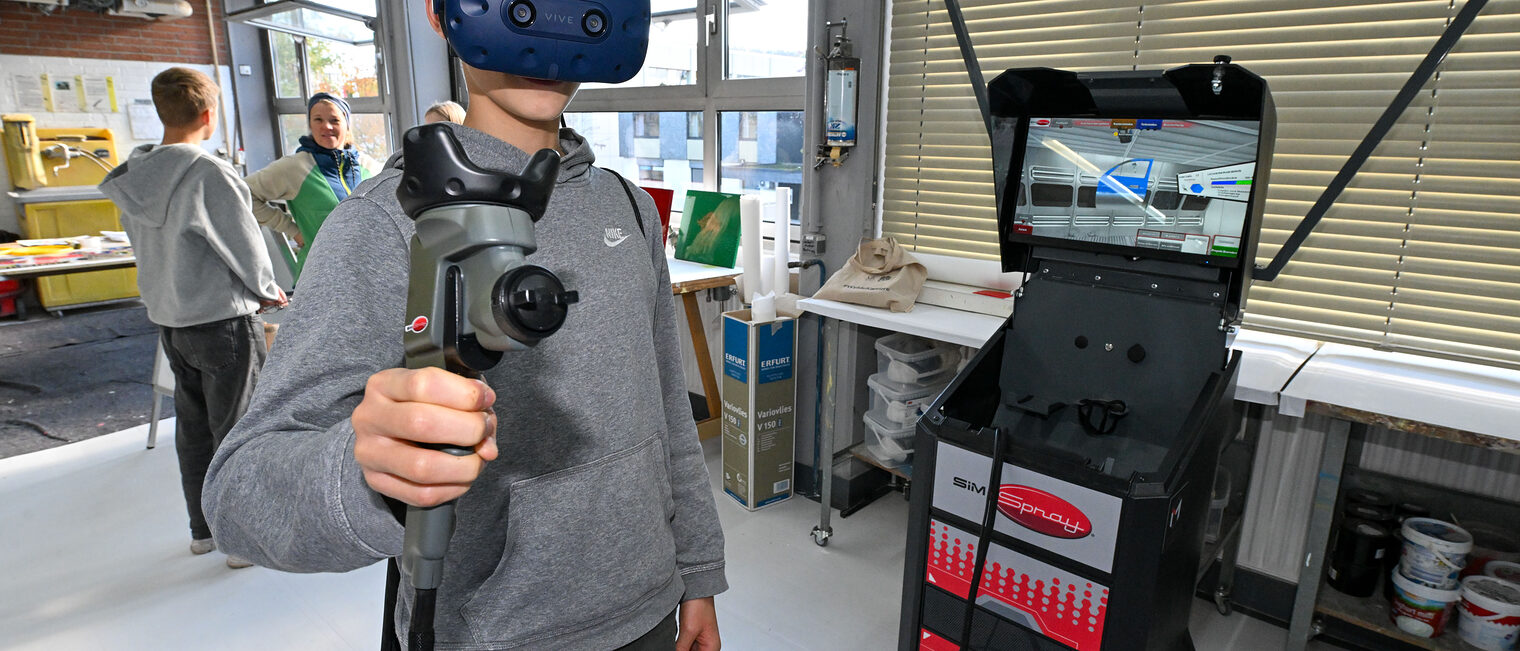 Ein Jugendlicher trägt eine VR-Brille und eine Spritzpistole und lackiert virtuell eine Autotür. 