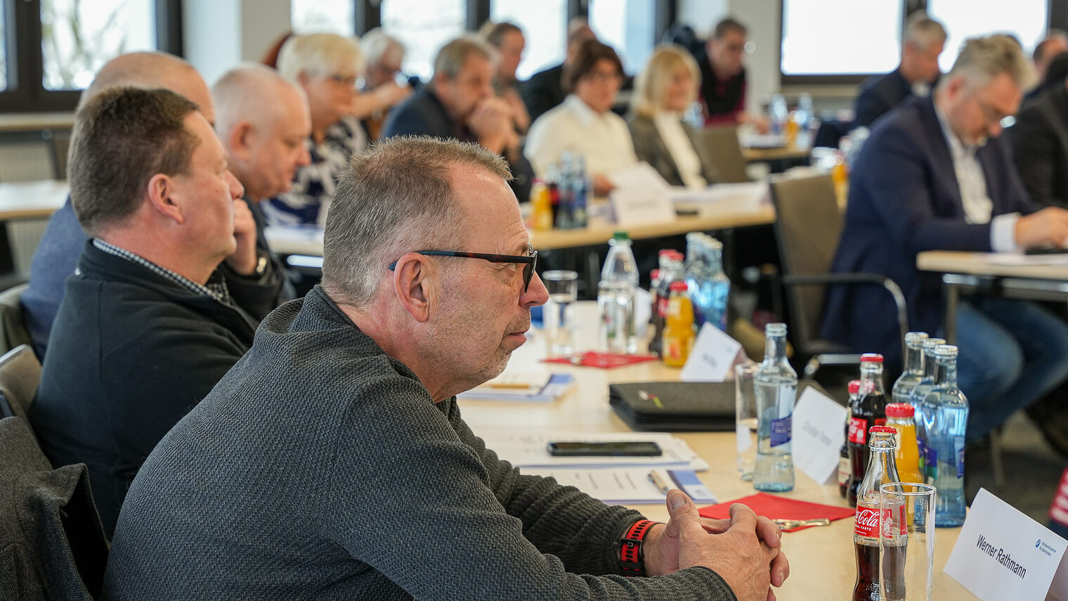 Mitglieder der Vollversammlung sitzen an Tischen und hören dem Vortragenden zu.