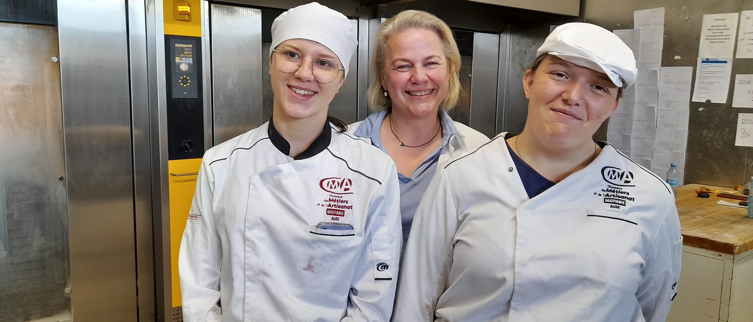 Zwei französische Auszubildende posieren mit einer Ausbildungsleiterin vor den Kühlräumen einer Bäckerei.