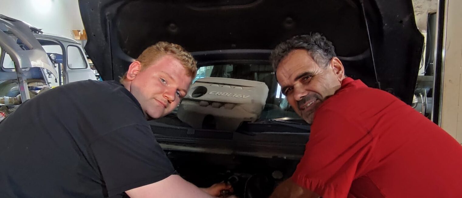 Ein deutscher Azubi und Ausbilder werkeln im Motorraum eines Autos in einer Werkstatt in Lezignan.