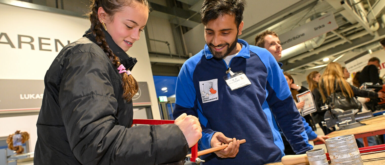 Eine Schülerin schneidet unter Anleitung ein Metallrohr mit einer Säge im Schraubstock zurecht.
