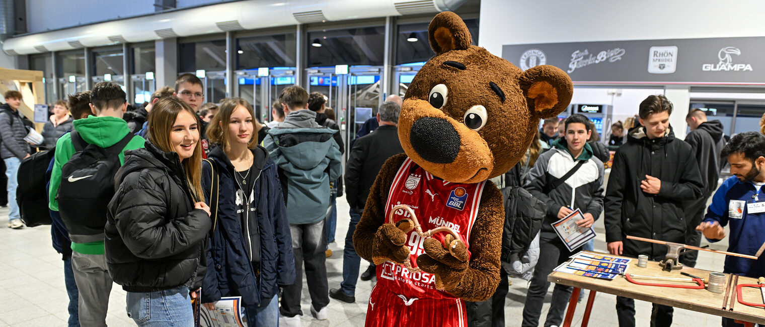 Maskottchen Freaky zeigt stolz ein Herz aus Kupferrohren, während Schülerinnen zusehen.