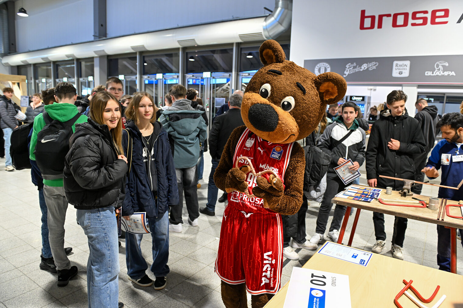 Maskottchen Freaky zeigt stolz ein Herz aus Kupferrohren, während Schülerinnen zusehen.